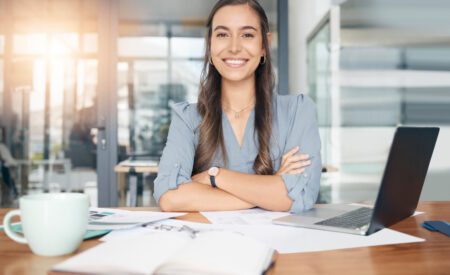 Business woman, portrait and smile at desk in office for paperwork, laptop or administration in Canada. Happy, young or confident female worker with pride planning project at table in startup company.